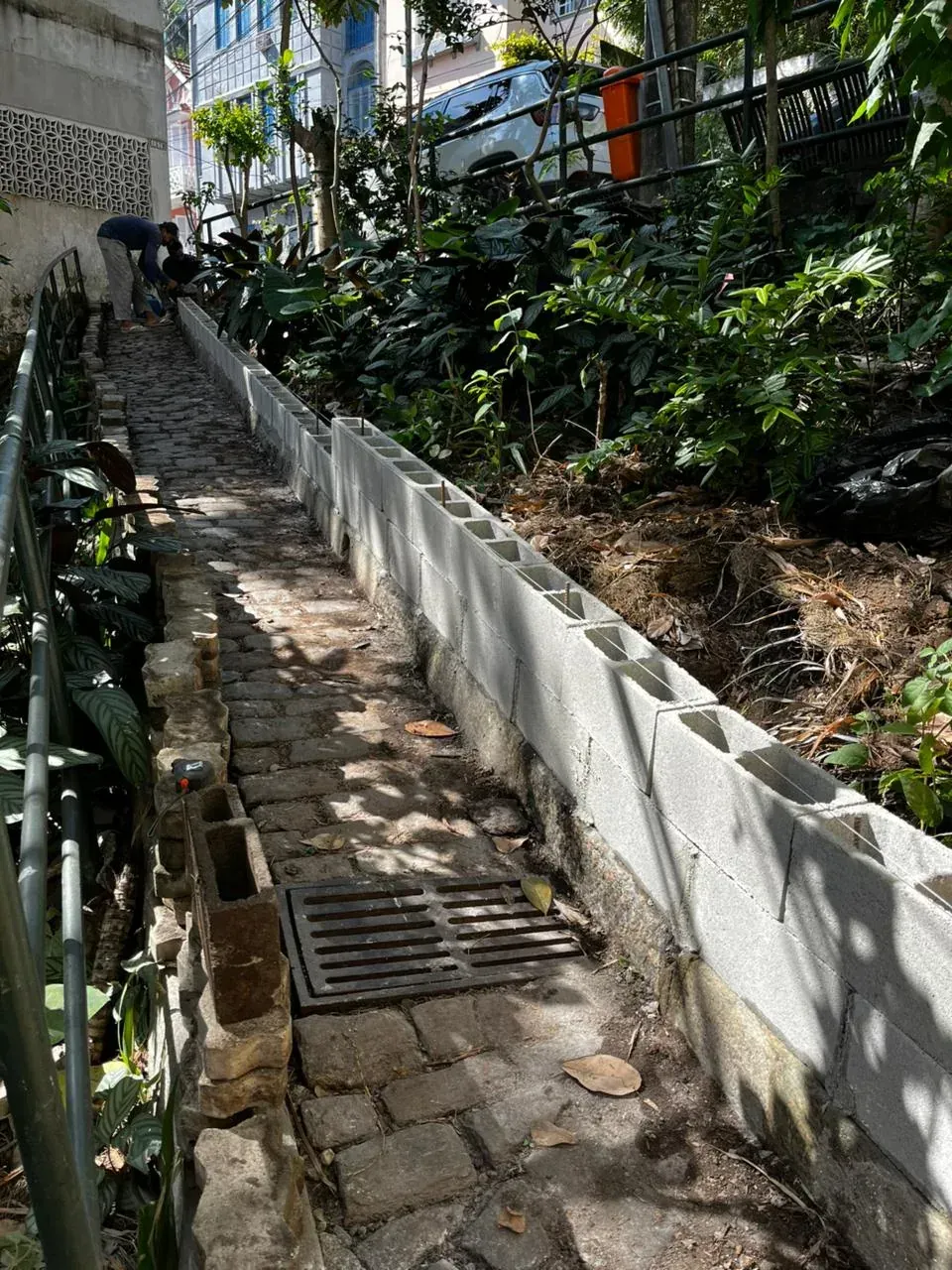 Muro de blocos de cimento recém-erguido ao longo do canteiro, com o calçamento de pedra da escadaria ao lado