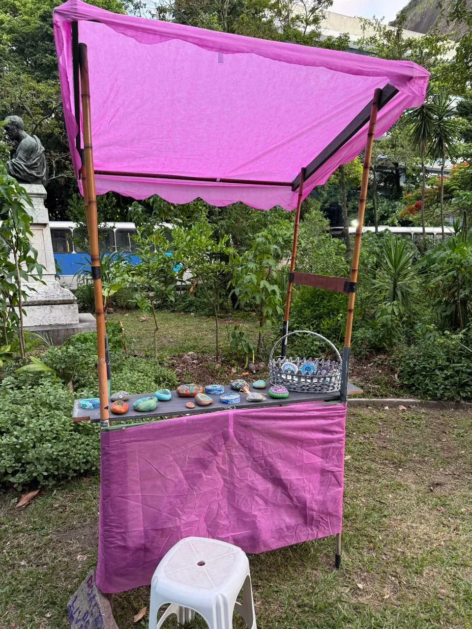 Barraca de feira com armação de bambu e cobertura de lona rosa, com pedras pintadas à mão expostas sobre a bancada, na praça arborizada