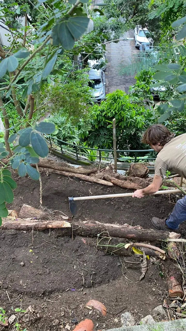 Canteiro em terraço de terra escura, com bordas feitas de troncos deitados, sendo aberto com enxada na encosta acima da rua