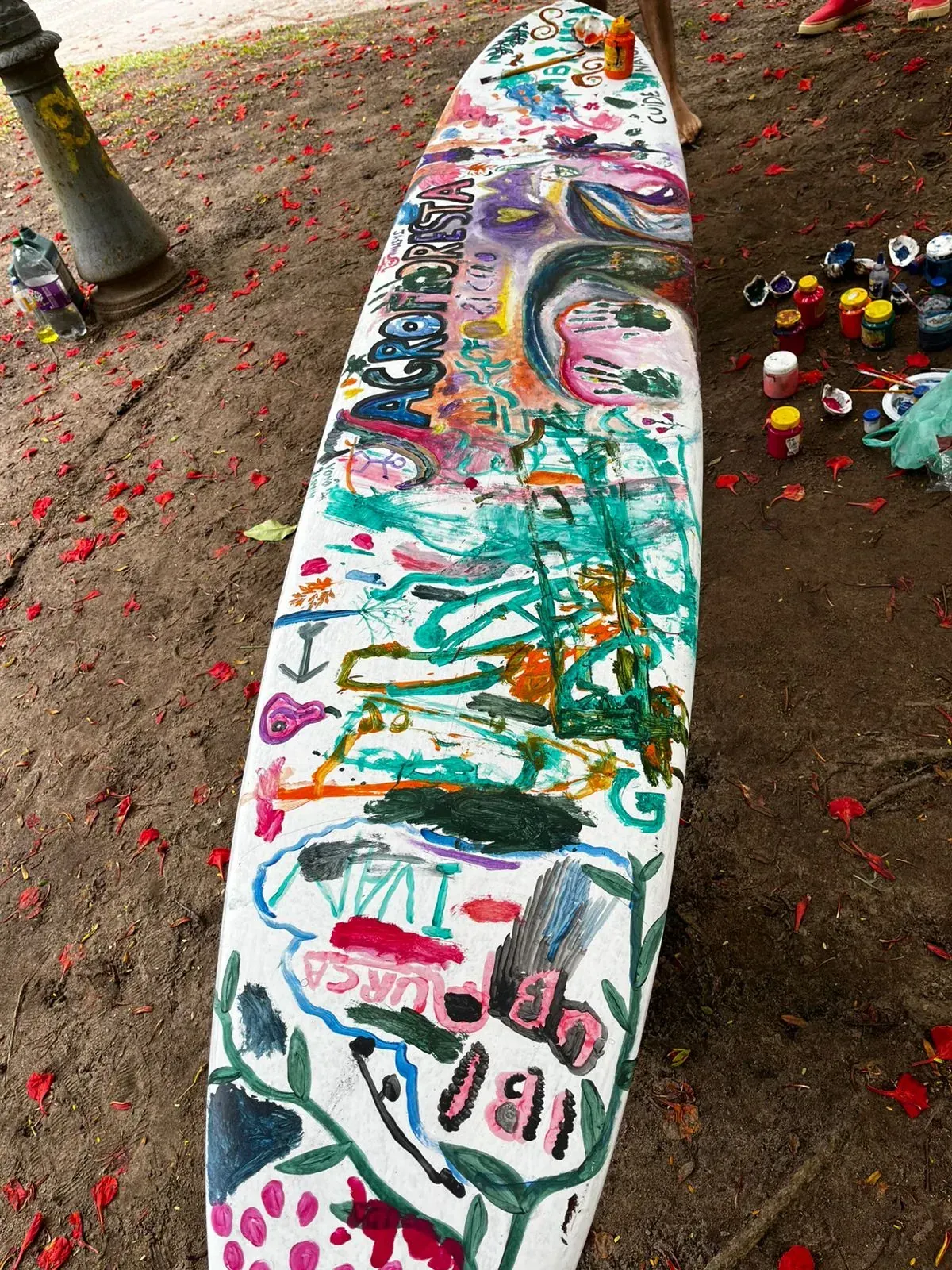 Prancha de stand-up deitada na terra coberta de flores vermelhas caídas, pintada à mão de ponta a ponta com plantas e a palavra AGROFLORESTA, cercada de potes de tinta